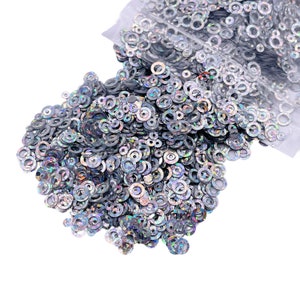 May include: A large pile of iridescent silver sequins in the shape of circles. The sequins are loose and spilling out of a clear plastic bag.