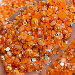 May include: A large assortment of orange and clear rhinestones and beads. The rhinestones are various sizes and shapes, including round, oval, and square. The beads are mostly round and have a pearlescent finish.