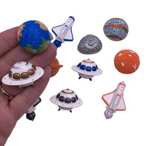 May include: A set of colorful space-themed figurines, including a blue and white planet, a silver asteroid, a white spaceship with a blue and white design, a white spaceship with a red and white design, a white flying saucer with a blue and white design, a white flying saucer with a black and white design, an orange planet, and a brown and orange planet.
