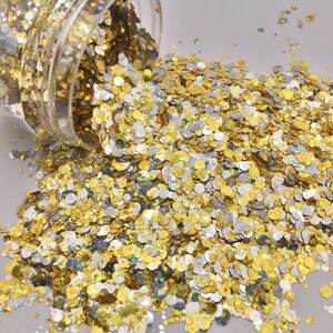 May include: A close-up of a jar of gold and silver glitter. The glitter is spilling out of the jar and is scattered on a white surface.