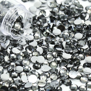 May include: A collection of gray, faceted rhinestones scattered on a white surface, with some spilling from a clear plastic container. The rhinestones have a flat back and a rounded, multi-faceted top, reflecting light. Ideal for crafts and embellishments.