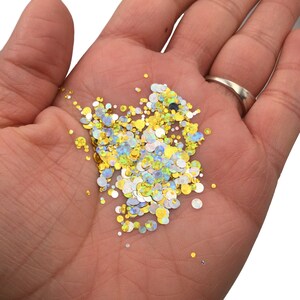 May include: A close-up of a hand holding a variety of iridescent glitter in gold, silver, and blue colors. The glitter is in the shape of small circles.