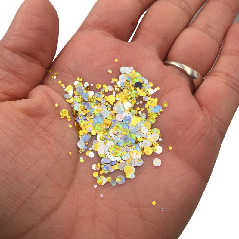 May include: A close-up of a hand holding a variety of iridescent glitter in gold, silver, and blue colors. The glitter is in the shape of small circles.