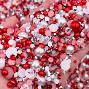 May include: A large assortment of red, white, and pink pearl and rhinestone embellishments. The embellishments are various sizes and shapes, including round, flat, and teardrop.