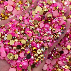 May include: A close-up of a hand holding a large assortment of pink and gold embellishments. The embellishments include small pearls, rhinestones, and flat-backed gems.