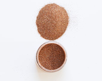 May include: A small jar of copper glitter with a pile of glitter spilled out next to it on a white surface.