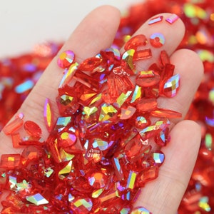 May include: A hand holding a variety of red iridescent rhinestones in different shapes and sizes.