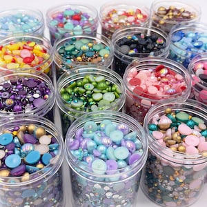 May include: Assortment of small containers filled with colorful beads and rhinestones. The containers are clear plastic and are arranged in rows. The beads are various shapes and sizes, including round, square, and star-shaped.