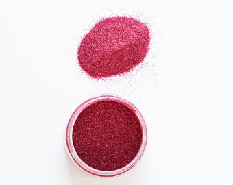 May include: A small jar of red glitter with a pile of red glitter next to it.