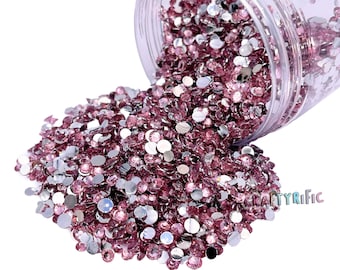 Pode incluir: Um recipiente de plástico transparente cheio de uma grande quantidade de pequenos strass redondos e iridescentes de cor rosa. O recipiente está tombado, derramando os strass sobre um fundo branco.