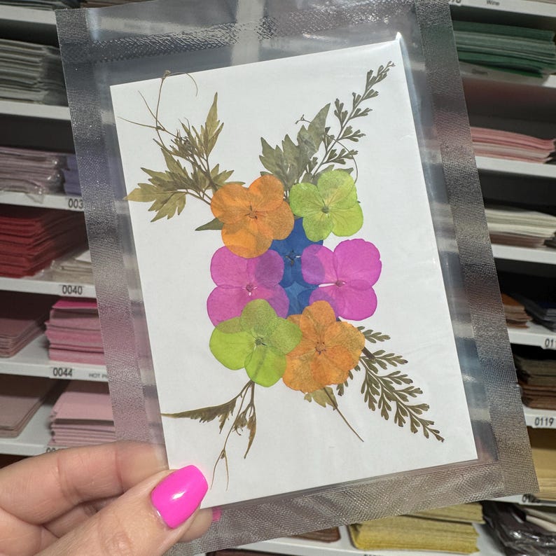 May include: A pressed flower art piece featuring a bouquet of colorful flowers, including pink, orange, green, and blue, with brown stems and leaves on a white background.