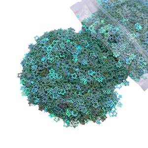 May include: A pile of iridescent green star-shaped glitter. The glitter is loose and scattered on a white background.