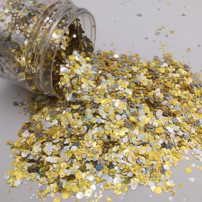 May include: A clear jar filled with gold and silver glitter, spilling out onto a white surface. The glitter is in the shape of small circles.