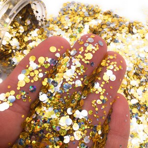 May include: A close-up of gold and silver glitter in a variety of sizes. The glitter is scattered on a white surface and is being held in a hand.