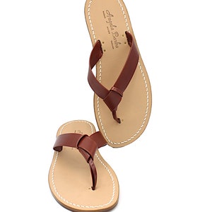 K&ouml;nnte beinhalten: Paar braune Leder-Flip-Flops mit einem geknoteten Riemendesign. Die Sandalen haben ein hellbraunes Fu&szlig;bett mit wei&szlig;en N&auml;hten an den R&auml;ndern. Der Markenname "Angela Barba" ist sichtbar.