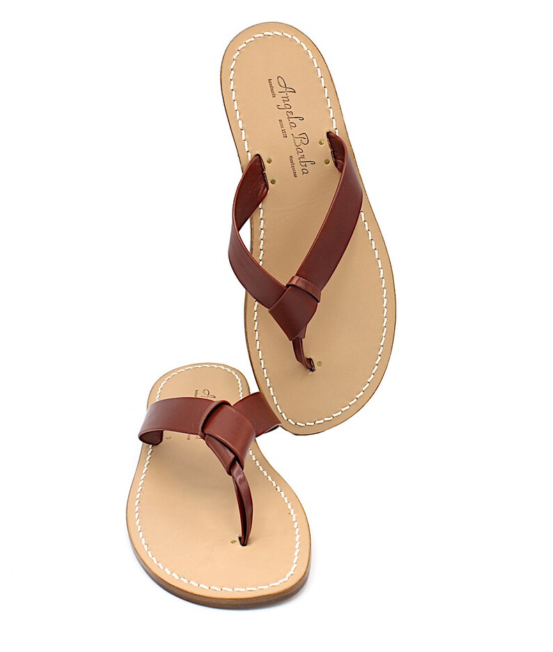 K&ouml;nnte beinhalten: Paar braune Leder-Flip-Flops mit einem geknoteten Riemendesign. Die Sandalen haben ein hellbraunes Fu&szlig;bett mit wei&szlig;en N&auml;hten an den R&auml;ndern. Der Markenname "Angela Barba" ist sichtbar.
