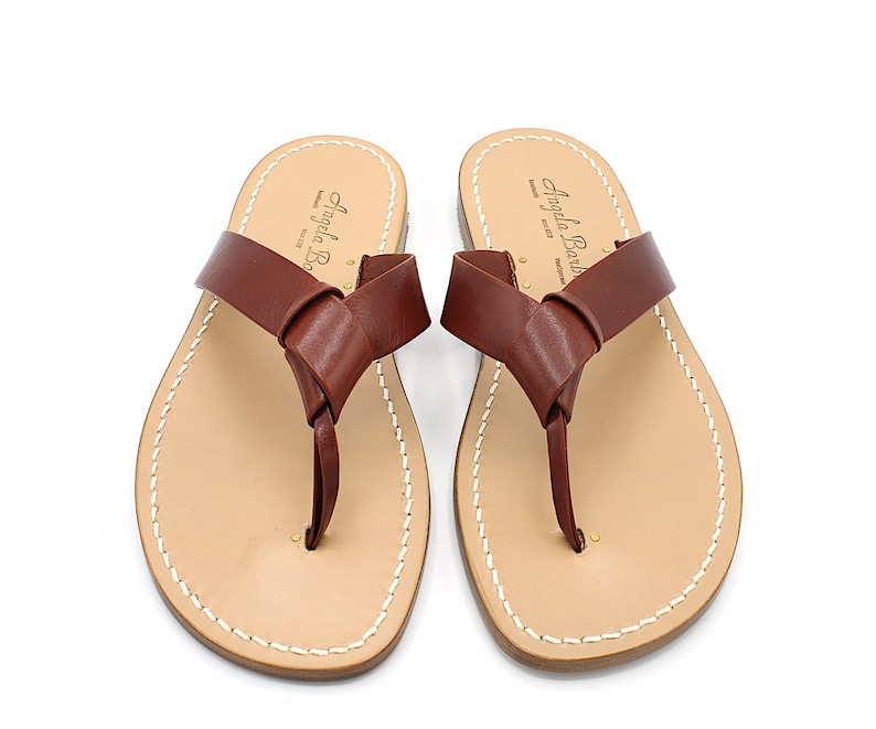 K&ouml;nnte beinhalten: Ein Paar braune Leder-Zehensandalen mit einem geknoteten Riemendesign. Die Sandalen haben ein hellbraunes Fu&szlig;bett mit wei&szlig;en N&auml;hten an den R&auml;ndern. Der Markenname "Angela Barb" ist auf dem Fu&szlig;bett aufgedruckt.