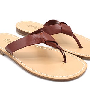 K&ouml;nnte beinhalten: Ein Paar braune Leder-Flip-Flops mit einem geknoteten Riemendesign. Die Sandalen haben ein hellbraunes Fu&szlig;bett mit wei&szlig;en N&auml;hten an den R&auml;ndern. Der Markenname "Angela B." ist auf dem Fu&szlig;bett aufgedruckt.