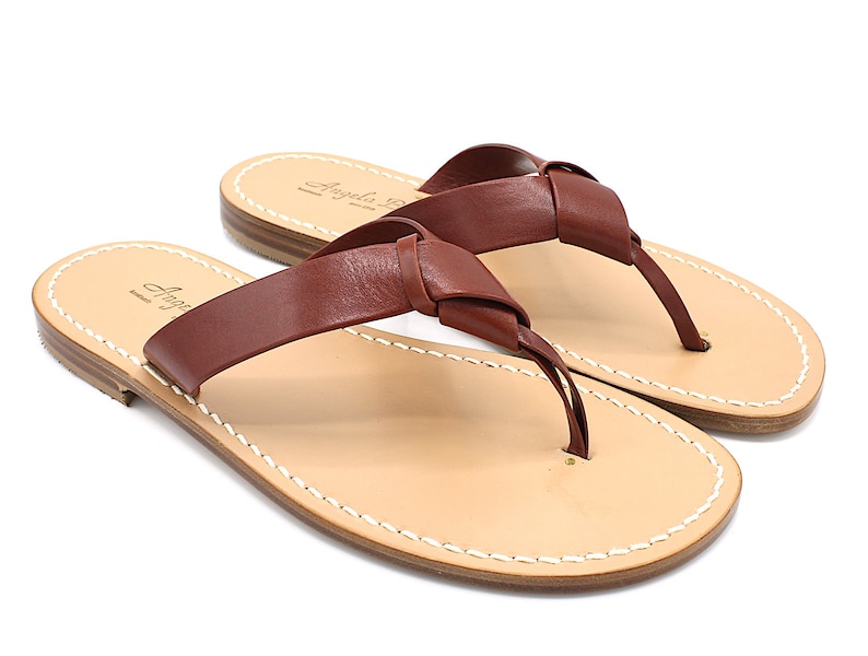 K&ouml;nnte beinhalten: Ein Paar braune Leder-Flip-Flops mit einem geknoteten Riemendesign. Die Sandalen haben ein hellbraunes Fu&szlig;bett mit wei&szlig;en N&auml;hten an den R&auml;ndern. Der Markenname "Angela B." ist auf dem Fu&szlig;bett aufgedruckt.