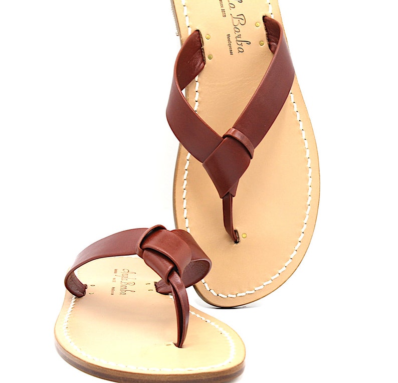 K&ouml;nnte beinhalten: Braune Ledersandalen mit einem Design mit geknoteten Riemen. Die Sandalen haben ein hellbraunes Fu&szlig;bett mit wei&szlig;en N&auml;hten an den R&auml;ndern. Der Markenname "Gigi Barba" ist auf dem Fu&szlig;bett aufgedruckt.