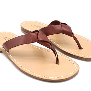 K&ouml;nnte beinhalten: Ein Paar braune Leder-Zehensandalen mit einem Design aus geknoteten Riemen. Die Sandalen haben ein hellbraunes Fu&szlig;bett mit wei&szlig;en N&auml;hten an den R&auml;ndern. Der Markenname "Angela" ist auf dem Fu&szlig;bett aufgedruckt.