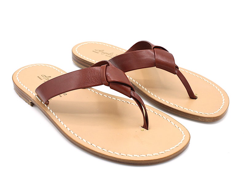 K&ouml;nnte beinhalten: Ein Paar braune Leder-Zehensandalen mit einem Design aus geknoteten Riemen. Die Sandalen haben ein hellbraunes Fu&szlig;bett mit wei&szlig;en N&auml;hten an den R&auml;ndern. Der Markenname "Angela" ist auf dem Fu&szlig;bett aufgedruckt.