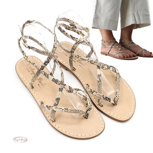 Könnte beinhalten: Ein Paar beigefarbene Ledersandalen mit Schlangenmuster. Die Sandalen haben mehrere Riemen, die sich über Fuß und Knöchel kreuzen. Die Sandalen sind von der Marke "Angela Barbi" und haben eine flache Sohle.