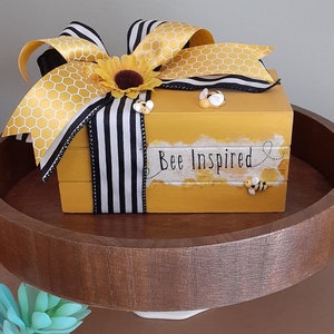May include: A yellow wooden box with a black and white striped ribbon and a honeycomb pattern bow. The box has the words "Bee Inspired" written on the front. There are small bee figurines on the box.