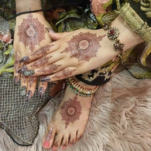 Op de afbeelding: Close-up van de handen en voeten van een persoon met ingewikkelde henna-ontwerpen. De henna is een diepe roodbruine kleur en bedekt de hele hand en voet. De persoon draagt een traditionele Indiase outfit en sieraden.