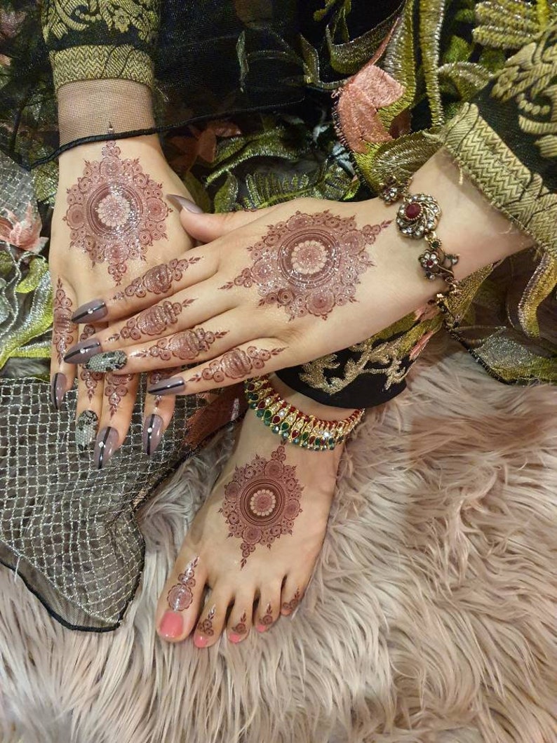 Op de afbeelding: Close-up van de handen en voeten van een persoon met ingewikkelde henna-ontwerpen. De henna is een diepe roodbruine kleur en bedekt de hele hand en voet. De persoon draagt een traditionele Indiase outfit en sieraden.