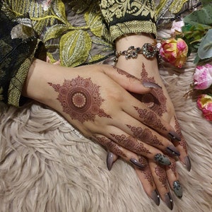 Op de afbeelding: Close-up van iemands handen met ingewikkelde henna-ontwerpen op de handpalmen en vingers. De henna is een diepe roodbruine kleur en de nagels zijn geverfd in een lichtgrijs met zwarte accenten. De persoon draagt een armband met een rode steen.