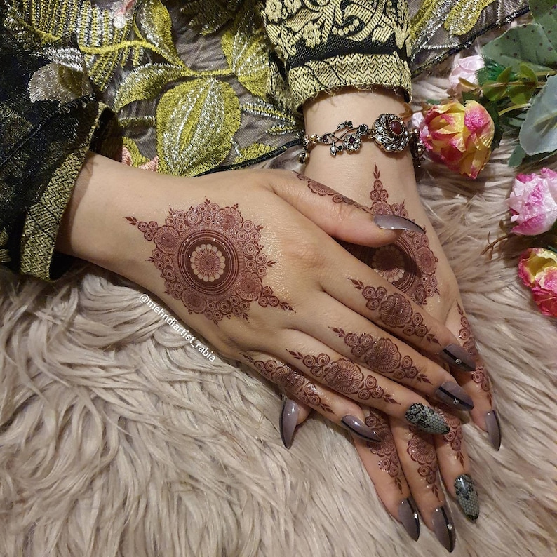 Op de afbeelding: Close-up van iemands handen met ingewikkelde henna-ontwerpen op de handpalmen en vingers. De henna is een diepe roodbruine kleur en de nagels zijn geverfd in een lichtgrijs met zwarte accenten. De persoon draagt een armband met een rode steen.