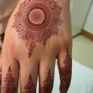 Op de afbeelding: Een hand met ingewikkelde henna-ontwerpen. De palm heeft een groot cirkelvormig ontwerp met kleinere cirkels en bladeren. De vingers hebben kleinere, symmetrische ontwerpen.