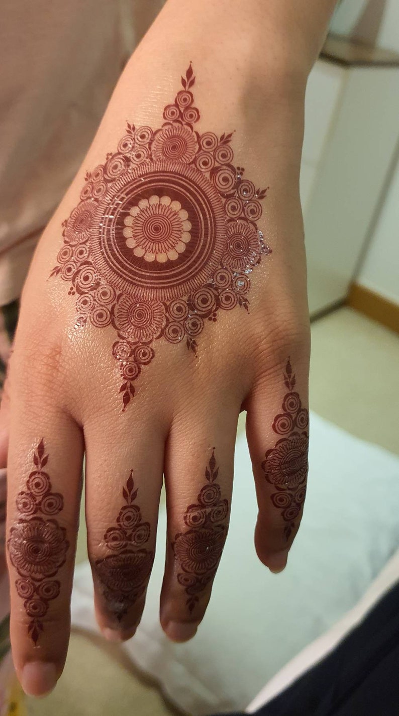Op de afbeelding: Een hand met ingewikkelde henna-ontwerpen. De palm heeft een groot cirkelvormig ontwerp met kleinere cirkels en bladeren. De vingers hebben kleinere, symmetrische ontwerpen.