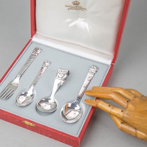 Può includere: Un set di quattro posate per bambini in argento sterling in una scatola rossa. Il set comprende una forchetta, due cucchiai e un coltello. Le posate sono decorate con un motivo floreale.