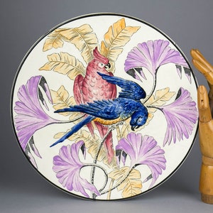 May include: A white ceramic plate featuring a hand-painted design of two parrots perched on a branch with purple flowers and yellow leaves.
