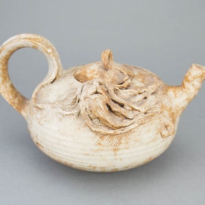 Peut inclure: Une théière en céramique blanche avec une surface texturée et un glaçage brun. La théière a un corps arrondi et un bec verseur sur le côté. Le couvercle de la théière a un motif en relief.