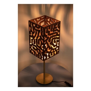 May include: A wooden table lamp with a geometric, abstract design. The lampshade is made of wood and has a light brown finish. The lamp base is gold metal.