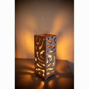 May include: A wooden, rectangular lamp with a carved, abstract design. The lamp is made of light-colored wood and has cut-out shapes that allow light to shine through. The lamp is lit from within, casting warm light and shadows.