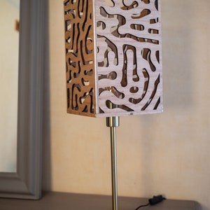May include: A gold table lamp with a wooden shade featuring a geometric pattern. The shade is made of thin wood with cutouts that create a unique design.