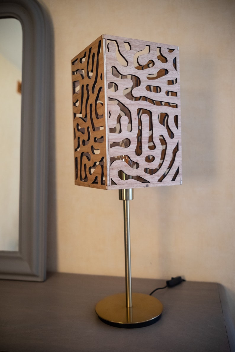 May include: A gold table lamp with a wooden shade featuring a geometric pattern. The shade is made of thin wood with cutouts that create a unique design.