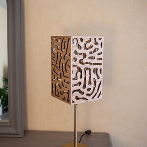 May include: A wooden table lamp with a geometric patterned shade. The shade is made of light brown wood with a cutout design. The lamp has a gold metal base and a cord.