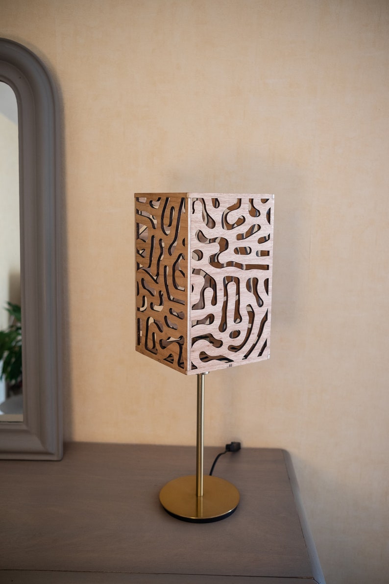 May include: A wooden table lamp with a geometric patterned shade. The shade is made of light brown wood with a cutout design. The lamp has a gold metal base and a cord.