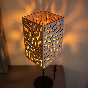 May include: A wooden table lamp with a geometric, abstract design. The lampshade is made of light wood with a cutout pattern and is illuminated from within. The lamp has a black metal base.