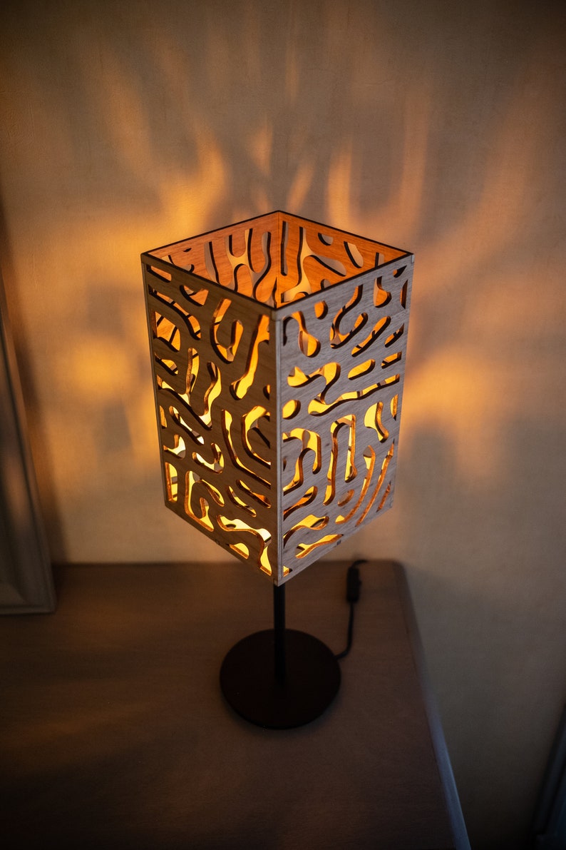 May include: A wooden table lamp with a geometric, abstract design. The lampshade is made of light wood with a cutout pattern and is illuminated from within. The lamp has a black metal base.