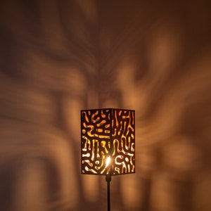 May include: A black table lamp with a wooden shade featuring a geometric pattern. The lamp is turned on and casting a warm glow.