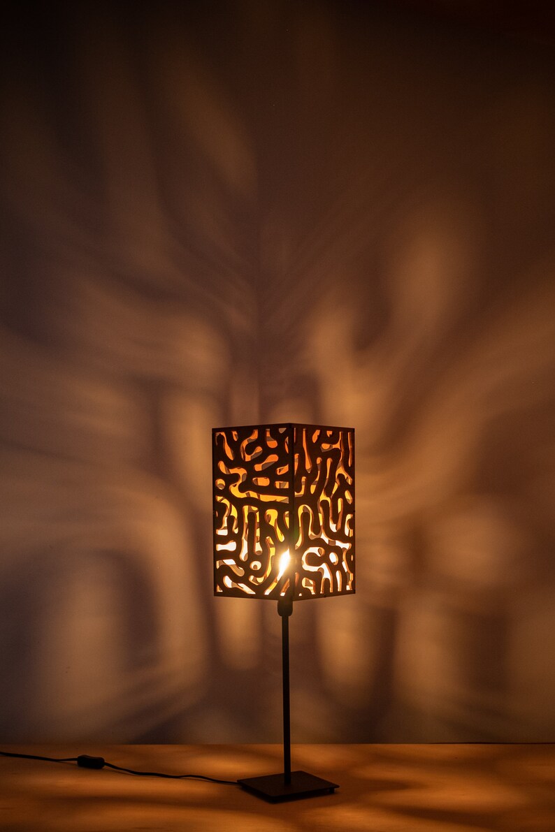 May include: A black table lamp with a wooden shade featuring a geometric pattern. The lamp is turned on and casting a warm glow.