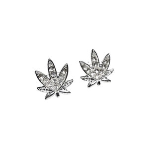 May include: A pair of silver-toned stud earrings in the shape of a cannabis leaf. Each leaf is encrusted with clear, sparkling crystals. The earrings are set against a plain white background, highlighting their intricate design and sparkle.