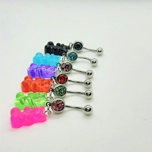 Può includere: Un set di sette anelli per l'ombelico con ciondoli a forma di orsacchiotto gommoso. I ciondoli sono di vari colori, tra cui rosa, verde, arancione, viola, blu, turchese e nero. Ogni anello ha un piccolo cristallo al centro.