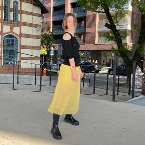 Puede incluir: Una mujer con un top negro con aberturas en los hombros y una falda larga de color amarillo brillante. Lleva botas negras. El fondo incluye un edificio de ladrillo y un árbol.